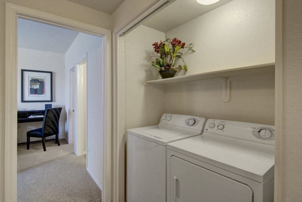 Laundry area here at Bela Rosa Apartments with a white washer and dryer, overhead shelf holding a plant with red flowers. Adjacent room features a desk, chair, and abstract artwork.