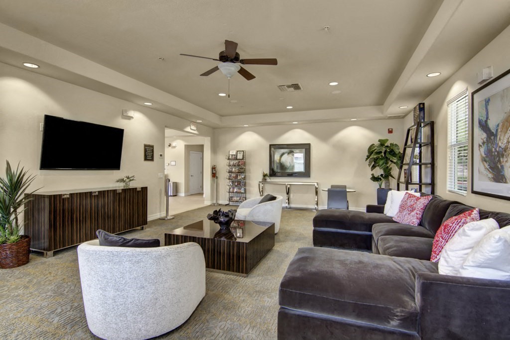 Modern residents' lounge here at Bela Rosa Apartments with neutral tones, featuring a large sectional sofa with red and white pillows, a flat-screen TV, and decorative plants. A cozy, inviting atmosphere is highlighted by soft lighting and contemporary artwork.