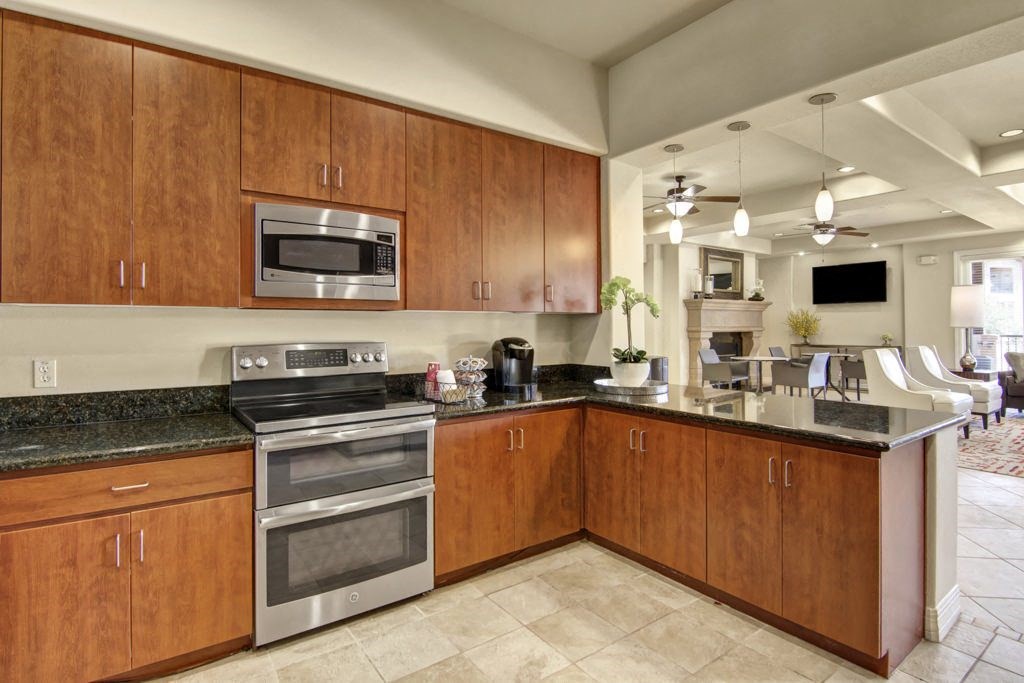 Modern kitchen here at Bela Rosa Apartments with wooden cabinets, black countertops, and stainless steel appliances. In the background, a bright dining area with pendant lights.