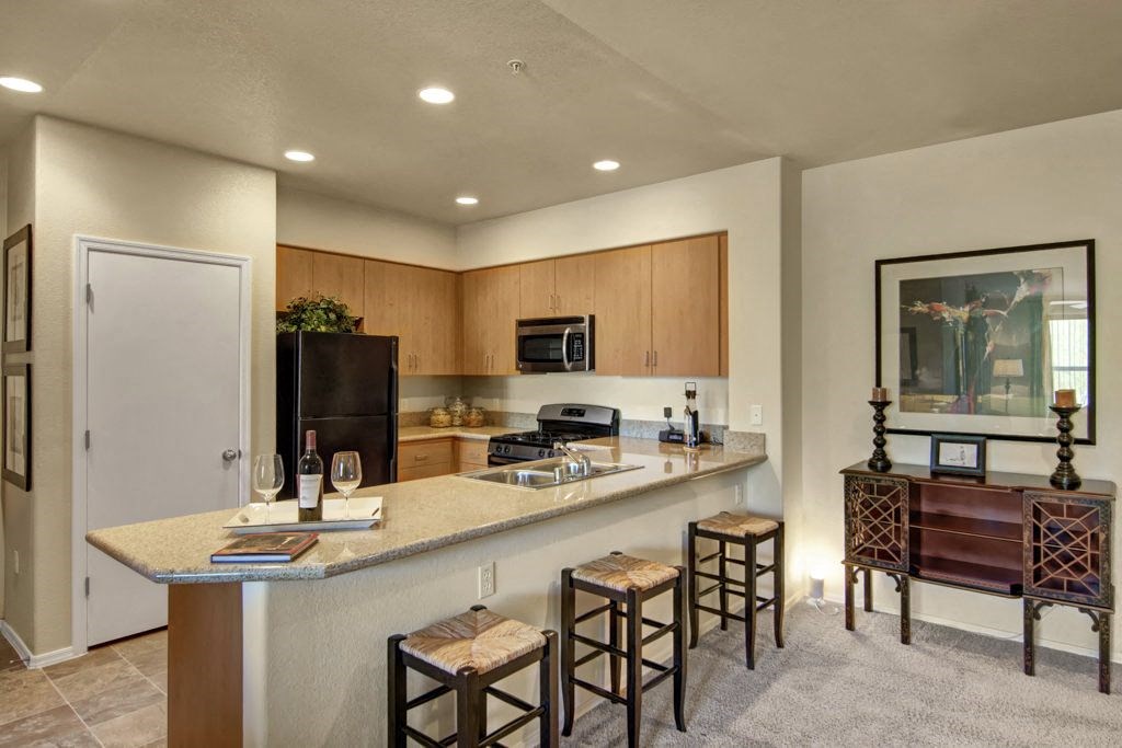 Modern kitchen here at Bela Rosa Apartments with wooden cabinets, black appliances, and a stone countertop. Three stools sit at the bar, with wine glasses and bottles, creating a cozy, inviting atmosphere.
