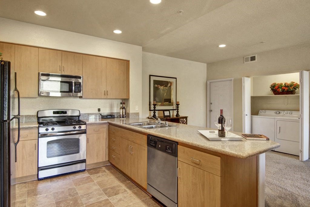 Modern kitchen here at Bela Rosa Apartments with light wood cabinets, stainless steel appliances, and granite countertops. Island holds a wine bottle and glass. Laundry room visible.