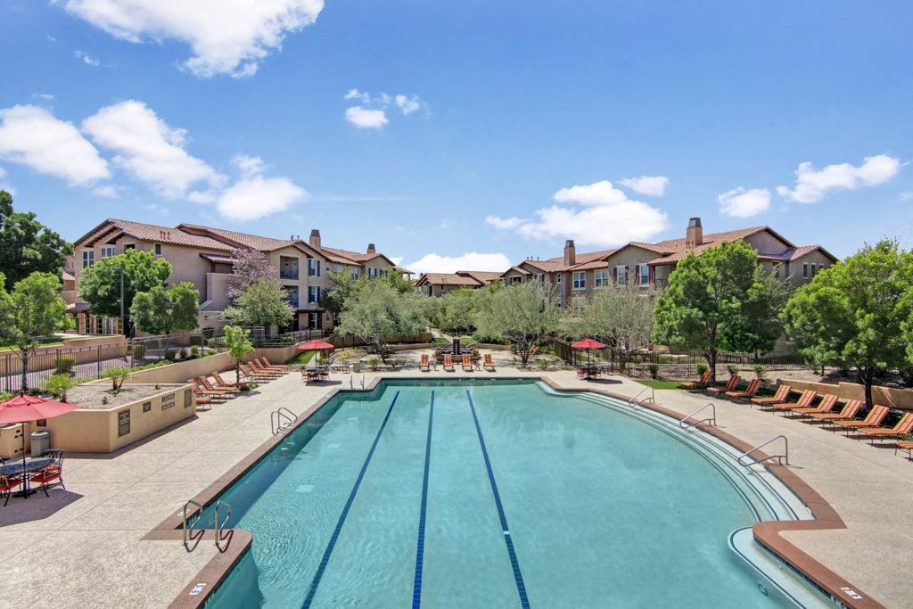 Outdoor swimming pool here at Bela Rosa Apartments surrounded by lounge chairs and umbrellas, with trees and apartment buildings in the background. The sky is clear and sunny.