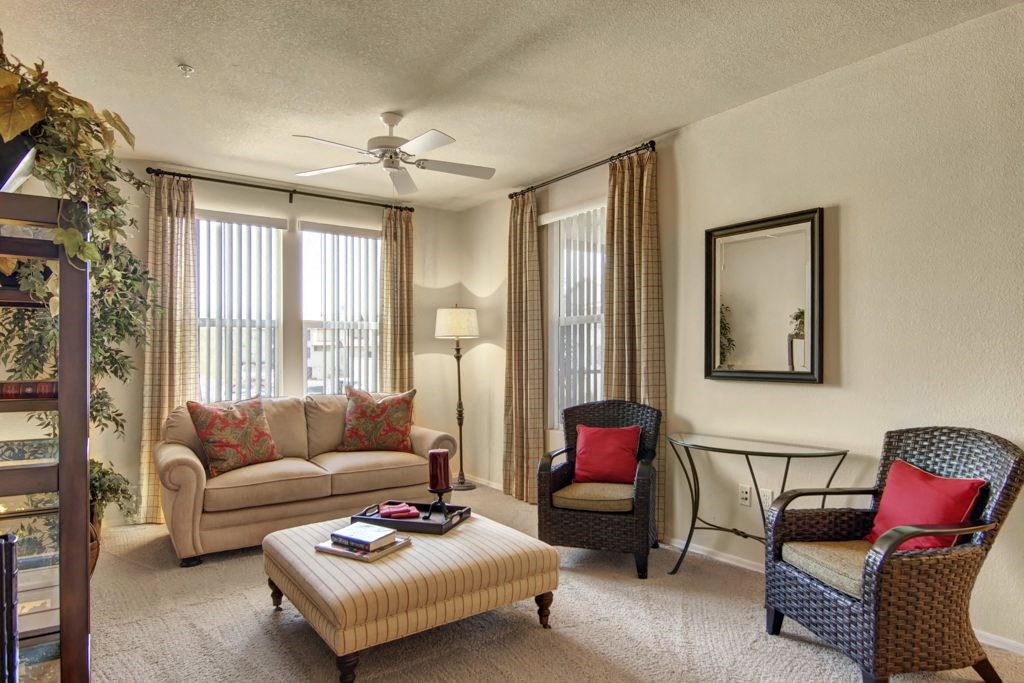 Cozy living room here at Bela Rosa Apartments with beige sofa, red-accented cushions, and two wicker chairs. Light streams through vertical blinds. Warm, inviting atmosphere.