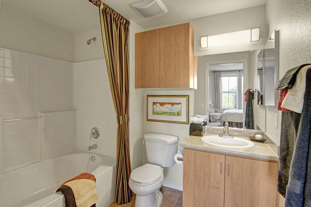 Compact bathroom here at Bela Rosa Apartments with white tub, striped curtain, wooden cabinets, and single sink. Bright lighting and cozy feel, opens to a bedroom via a doorway.
