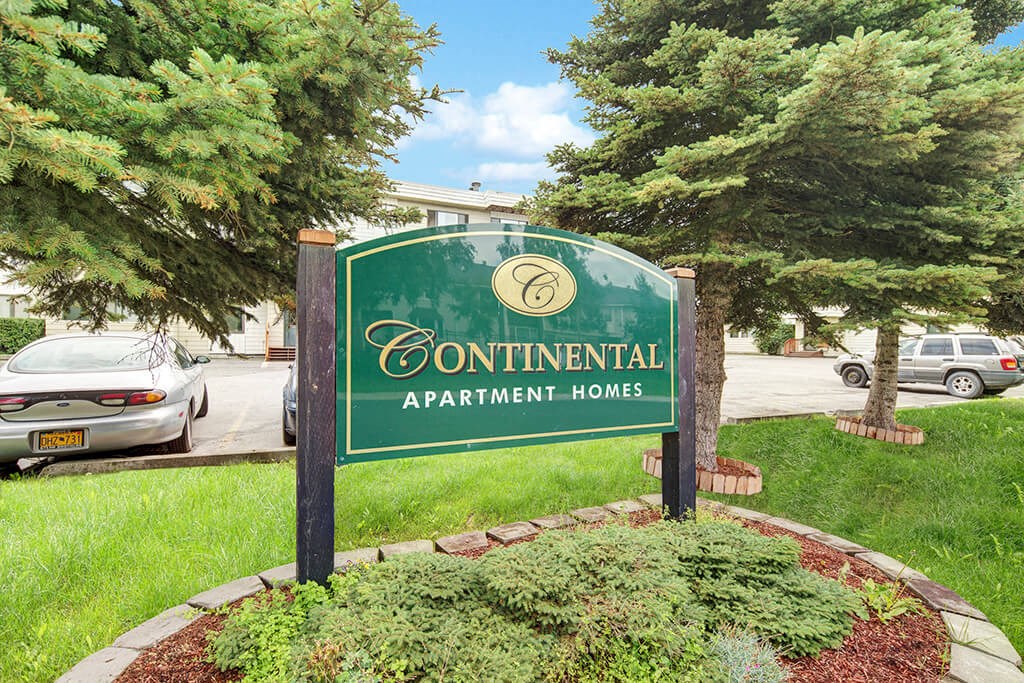 Sign for "Continental Apartment Homes" in a landscaped area with evergreen trees. Parked cars are visible in the background under a clear sky.