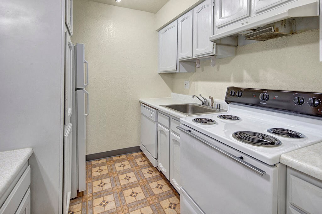 Continental Apartments - Kitchen Narrow kitchen here at The Continental Apartments with white cabinets, electric stove, sink, and refrigerator. Beige walls and patterned vinyl flooring add a simple, clean look.