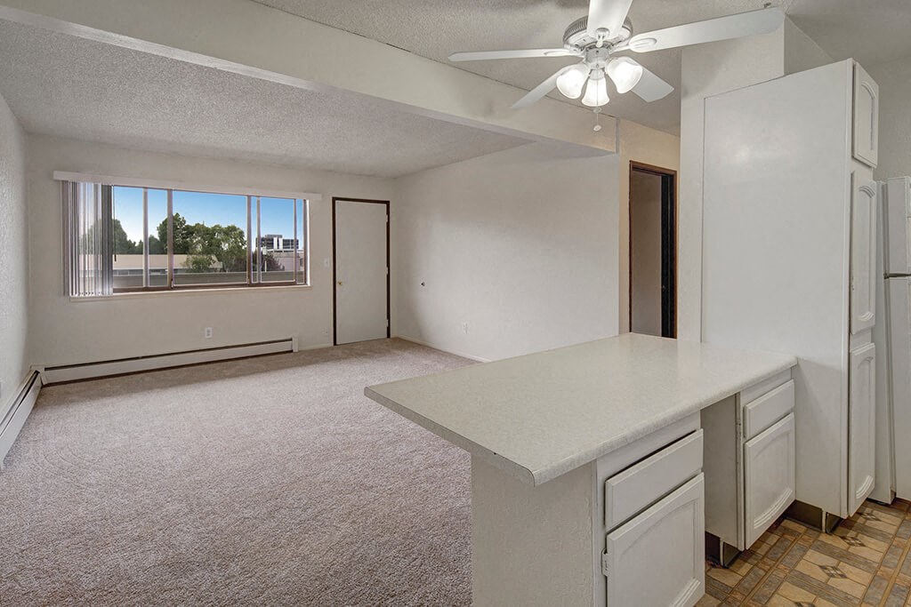 Spacious, empty living room here at The Continental Apartments with beige carpet, white walls, and large window. Kitchen with white cabinets and counter; ceiling fan present. Bright, airy feel.