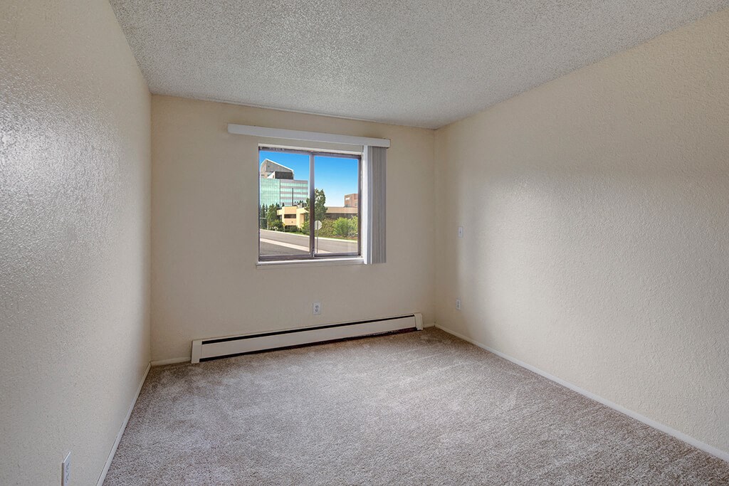 Continental Apartments - Bedroom Empty beige bedroom here at The Continental Apartments with textured walls and carpeted floor. A window reveals a bright, sunny view of modern buildings and greenery outside.