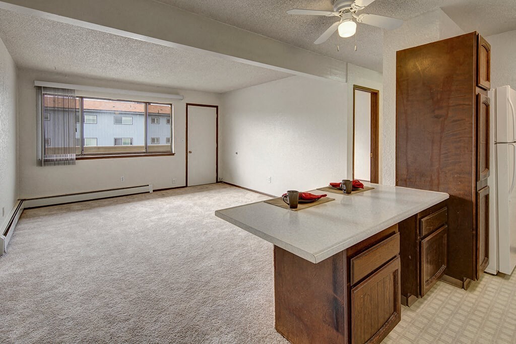 Continental Apartments - Living Room Spacious, empty living room here at The Continental Apartments with beige carpet and white walls. Large window with vertical blinds, overlooking adjacent buildings. Open kitchen counter with two place settings.
