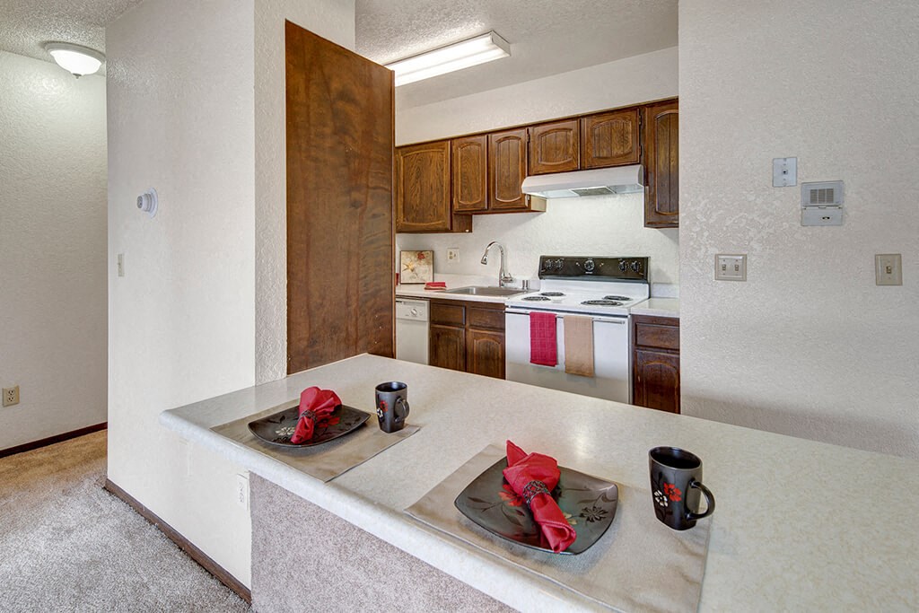 A cozy kitchen here at The Continental Apartments with wooden cabinets and white appliances. A countertop features two place settings with black dishes, red napkins, and mugs, creating a warm, inviting atmosphere.