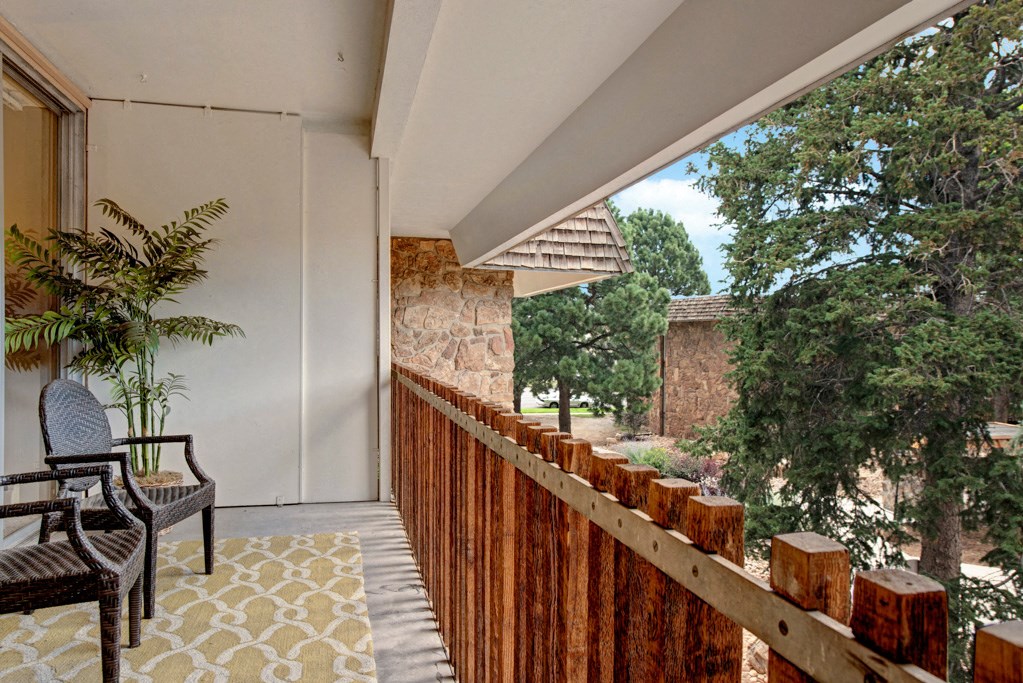 A cozy balcony here at Copper Stone Apartment Homes with wicker chairs, a potted plant, and a patterned rug overlooks a lush garden with tall trees and stone buildings under a cloudy sky.