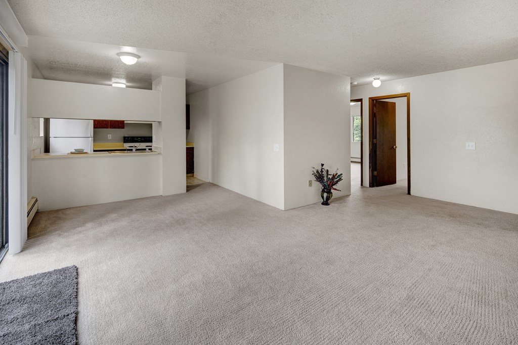 Spacious, empty living room here at Deer Park Apartments with light beige carpet, white walls, and a small vase with dried flowers. Kitchen with white appliances in the background.