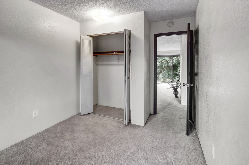 A carpeted bedroom here at Deer Park Apartments featuring an open closet and a doorway leading to a light-filled space with a window and potted plant.