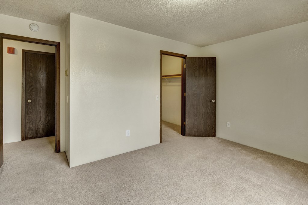 A spacious, empty room here at Deer Park Apartments with light beige carpet and neutral walls, featuring two closed wooden doors leading to closets.