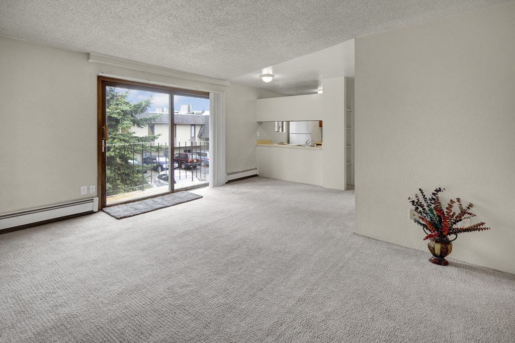Spacious, well-lit living room here at Deer Park Apartments with beige carpet, sliding glass doors leading to balcony, and an adjacent kitchen. A vase with colorful branches sits in the corner.