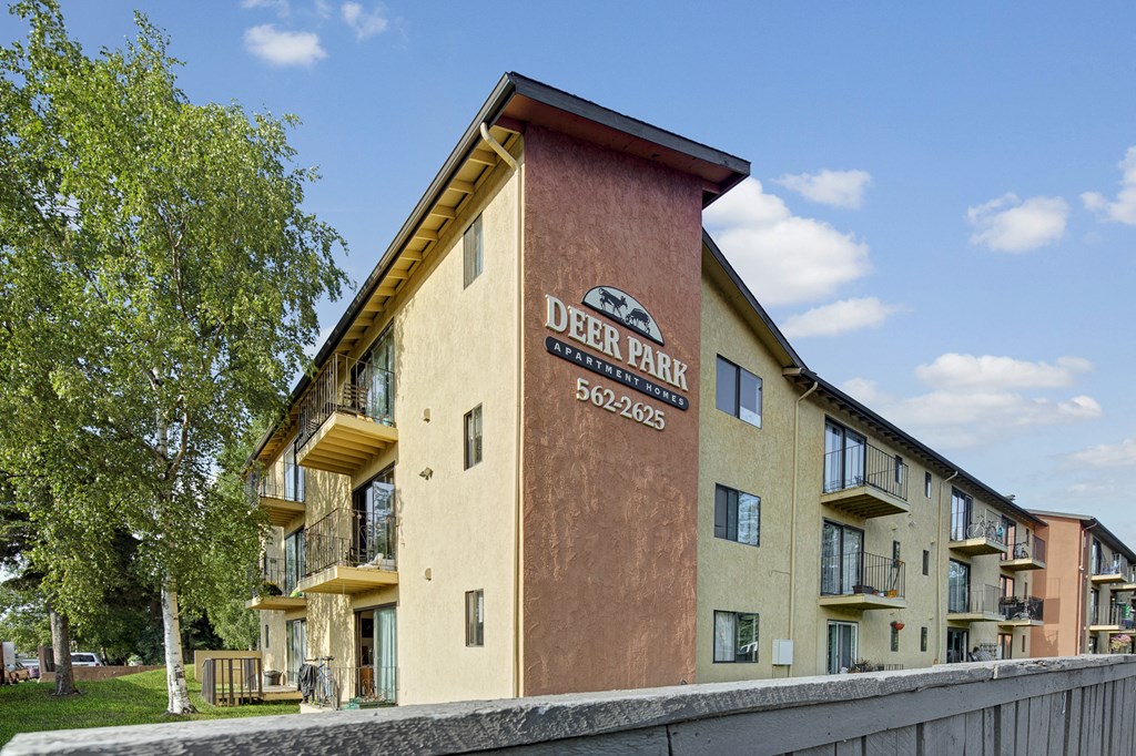 Exterior view of Deer Park Apartment Homes with balconies, a sign displaying the name and contact number, surrounded by greenery and blue sky.