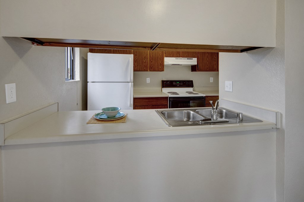 Minimalist kitchen here at Deer Park Apartments with a clean white countertop and a double sink, stainless steel faucet, white refrigerator, stove, wood cabinets, and a neatly placed blue dish set.