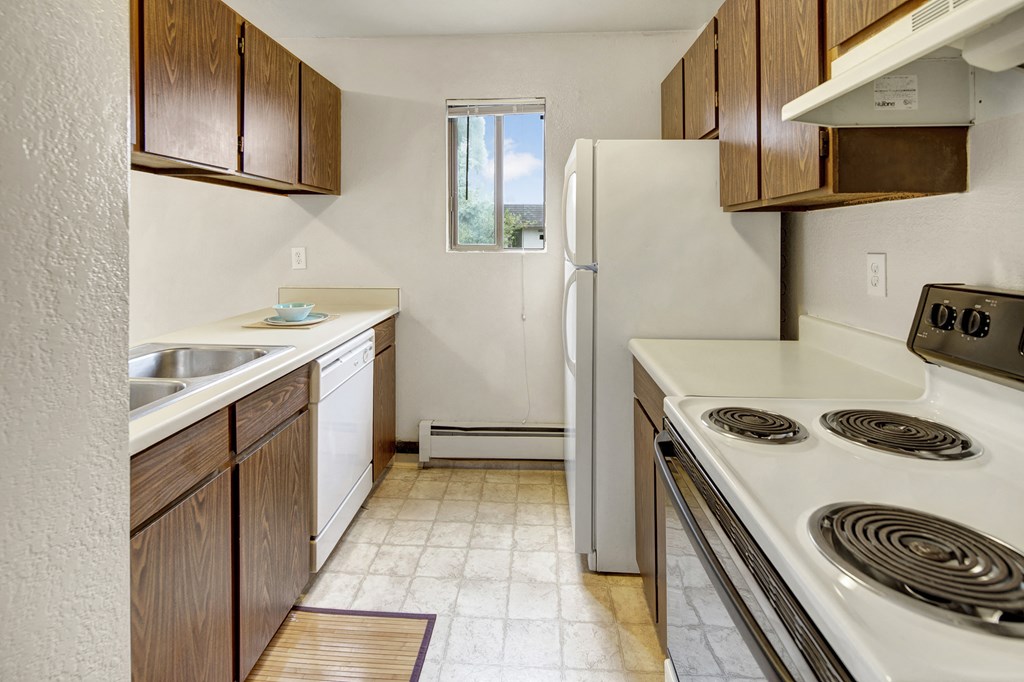 Compact kitchen here at Deer Park Apartments with wood cabinets, white appliances, and a small window. Includes a stove, double sink, and dishwasher. Bright, clean, and orderly.