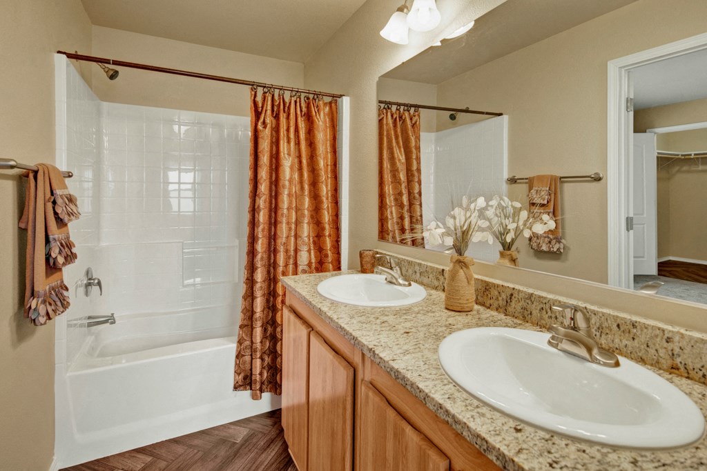 Bathroom here at Four Seasons Apartments with granite countertop, double sinks, and decorative vases. A white tub-shower with an orange curtain is in the background, conveying warmth.
