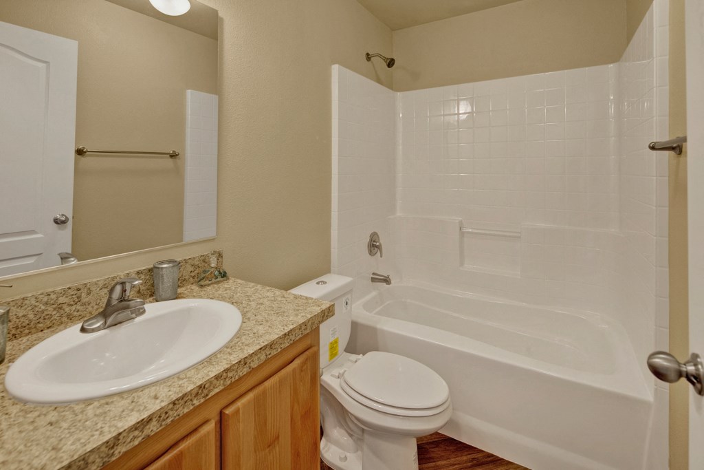Compact bathroom here at Four Seasons Apartments with a white bathtub-shower combo, granite countertop with sink, and wooden cabinetry. Neutral tones and simple, modern fixtures.