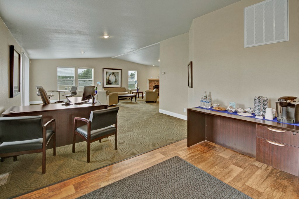 Office reception area here at Four Seasons Apartments with modern decor. Dark wood desks and chairs on green carpet. Refreshment station on the right. Bright, welcoming atmosphere.