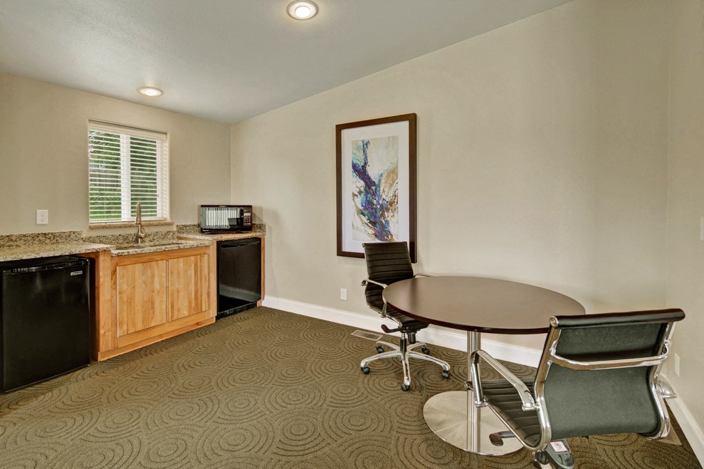 Minimalist office space here at Four Seasons Apartments with a round wooden table, two sleek black chairs, and a kitchenette with light wood cabinets. A colorful abstract painting adorns the beige wall.