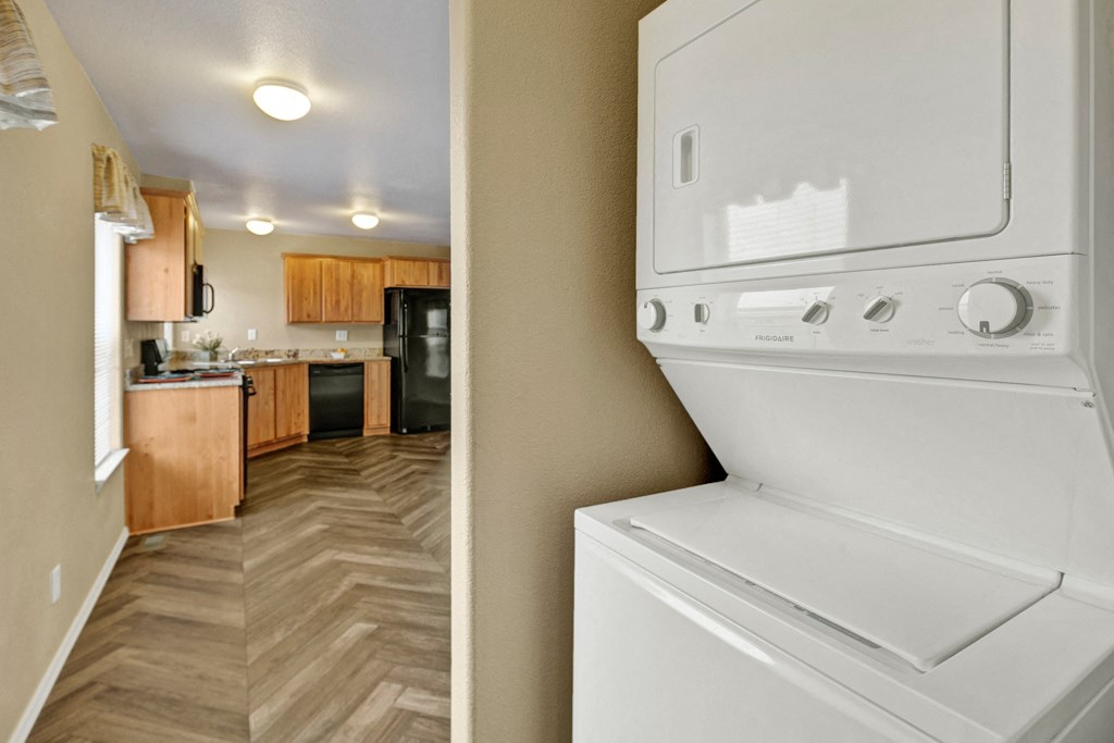 Stacked white washer and dryer in a hallway with a view of a kitchen. The kitchen here at Four Seasons Apartments features wooden cabinets, chevron-patterned flooring, and black appliances, conveying a cozy, modern feel.
