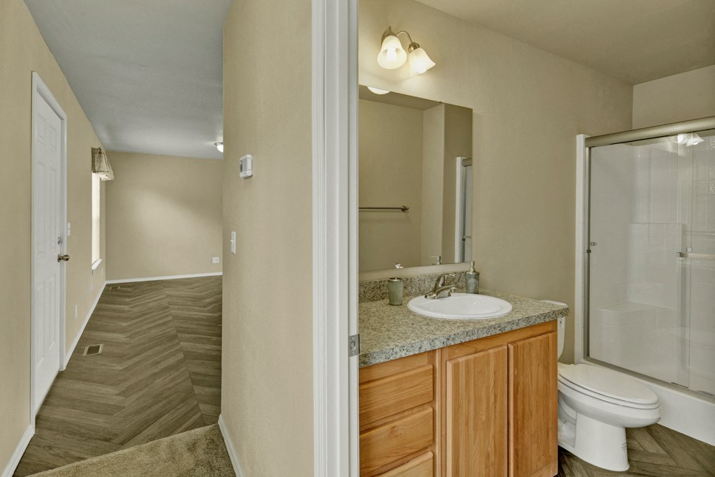 Hallway here at Four Seasons Apartments with wood-patterned floor leading to bathroom. Bathroom has a granite-topped wooden vanity, oval mirror, and shower. Walls are beige, creating a calm atmosphere.