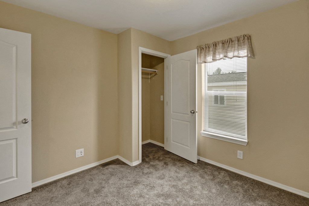 Beige bedroom here at Four Seasons Apartments with plush gray carpet, open closet, and window with blinds. Sunlight brightens the space, creating a calm, inviting atmosphere.