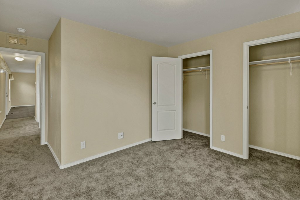 Bedroom here at Four Seasons Apartments with beige walls, open closet doors, and gray carpet. A hallway is visible leading to another lit room, creating a neutral and vacant atmosphere.
