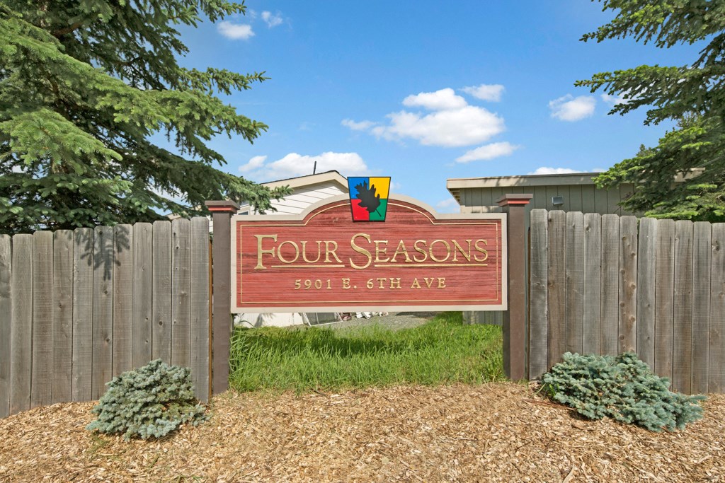 Wooden sign reading "Four Seasons, 5901 E. 6th Ave" surrounded by a wooden fence, green shrubs, and pine trees under a clear blue sky.