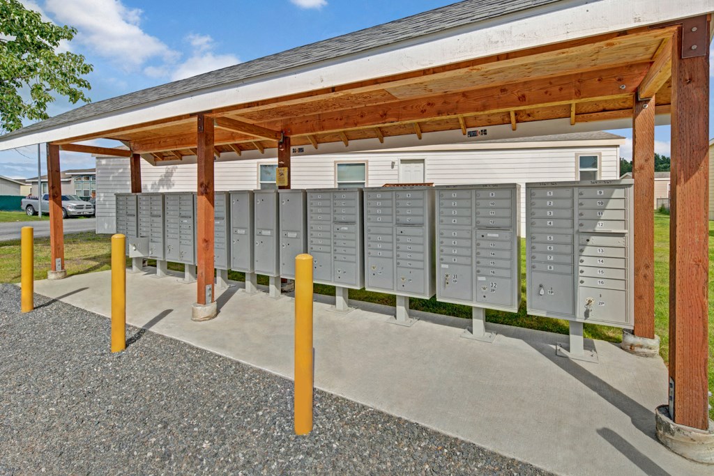 A covered outdoor mail station here at Four Seasons Apartments with multiple gray mailboxes lined up under a wooden shelter. Yellow bollards are in front on a gravel surface.