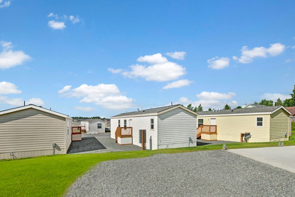 A row of mobile homes here at  Four Seasons Apartments with wooden decks is set on a neatly maintained grassy and gravel path under a bright blue sky with scattered clouds.