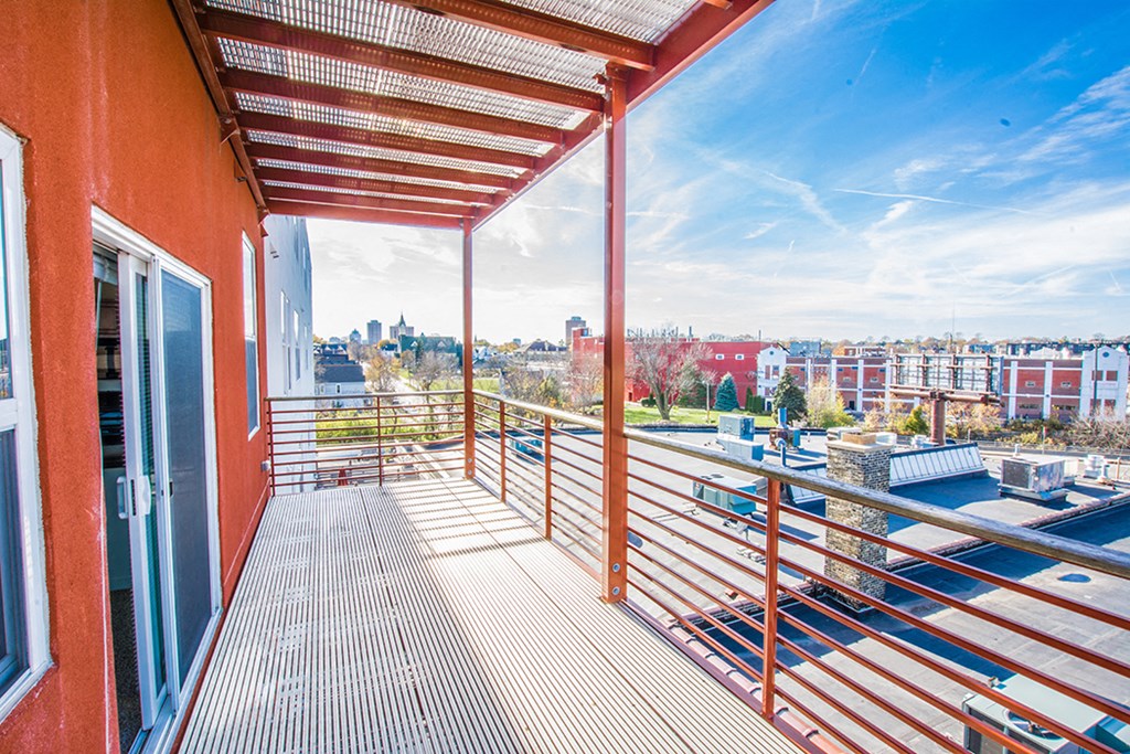Rooftop balcony at Latitude Apartments with red railing and wooden floor, overlooking an urban landscape with trees, buildings, and a clear blue sky. Bright, airy atmosphere.