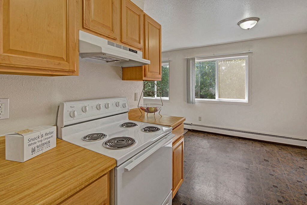 The Glen Apartments - Kitchen Simple kitchen here at The Glen Apartments with light wood cabinetry, white stove, and box labeled “Snack & Relax.” Bright window, empty fruit bowl, and clean, minimalist decor.