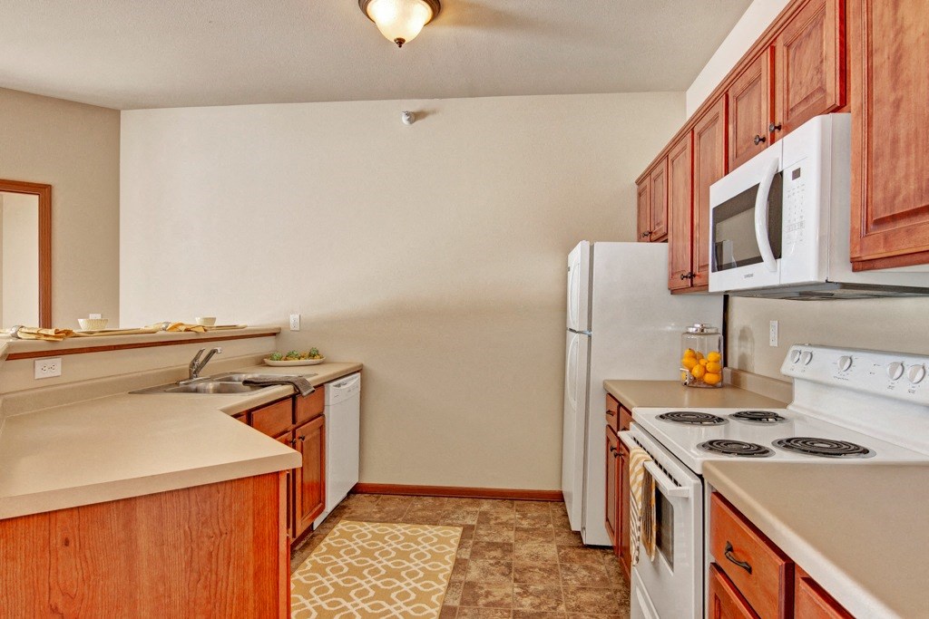 Warm kitchen here at Grand River Estates Apartment Homes with wood cabinets, white appliances, and beige countertops. A jar of oranges and neatly arranged dishware add a cozy, inviting feel.