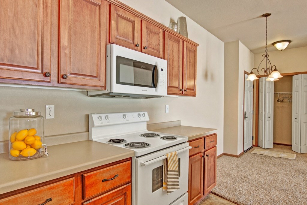 A cozy kitchen here at Grand River Estates Apartment Homes with wooden cabinets, a white stove, and a microwave. Lemons in a jar add a bright touch. Warm lighting sets an inviting tone.
