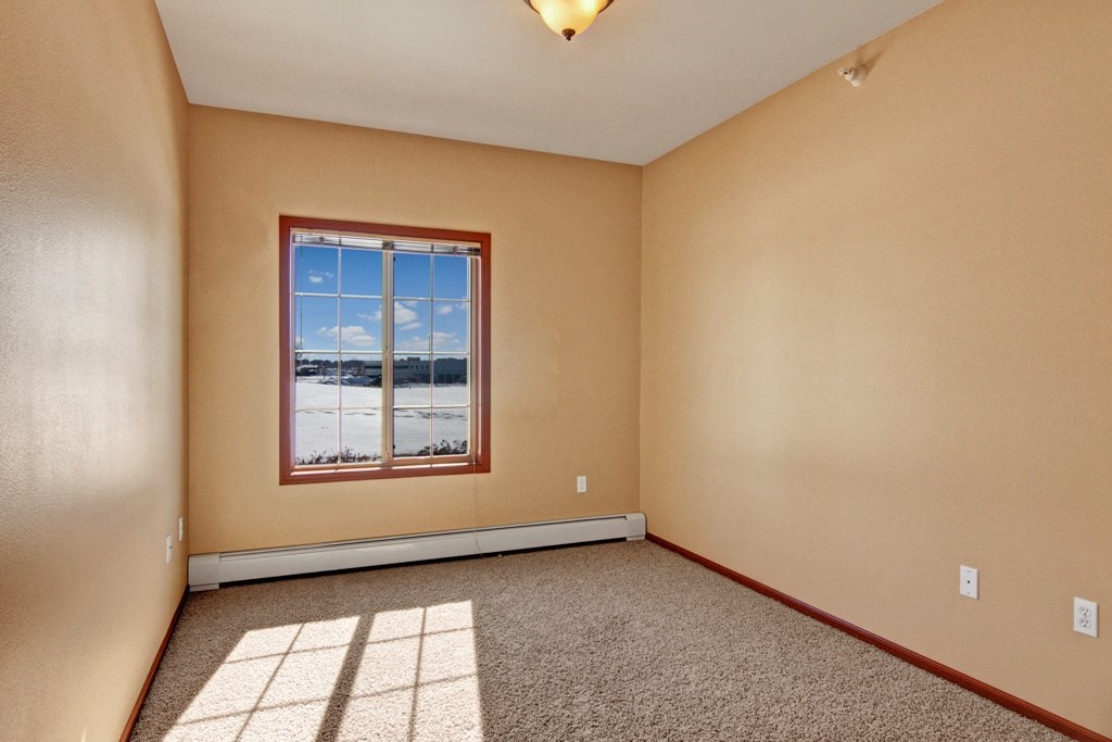 Empty room here at Grand River Estates Apartment Homes with beige walls and carpet, featuring a large window framing a snowy landscape and blue sky. The atmosphere is calm and serene.