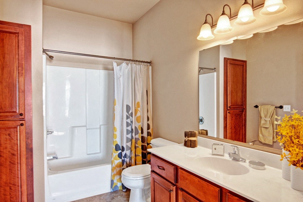 Bright bathroom here at Grand River Estates Apartment with wooden cabinets, a white counter, and double sink. Yellow accents include a vibrant shower curtain and flowers, under soft lighting.
