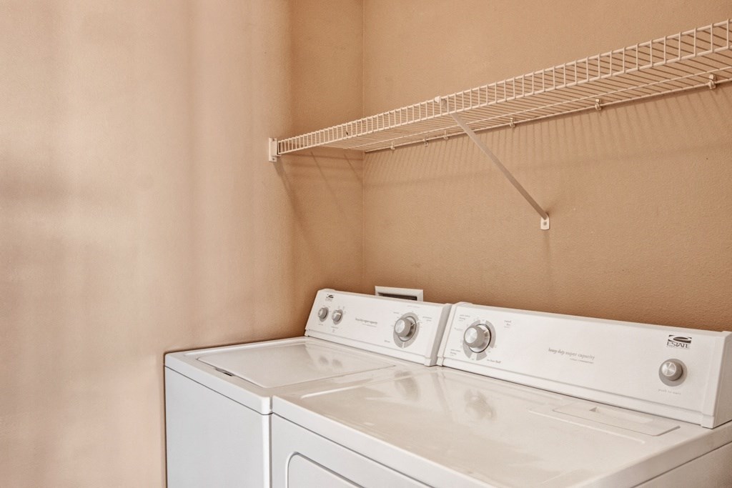 A bright laundry room here at Grand River Estates Apartment Homes with a white washer and dryer side by side against a beige wall. An empty white wire shelf hangs above them.