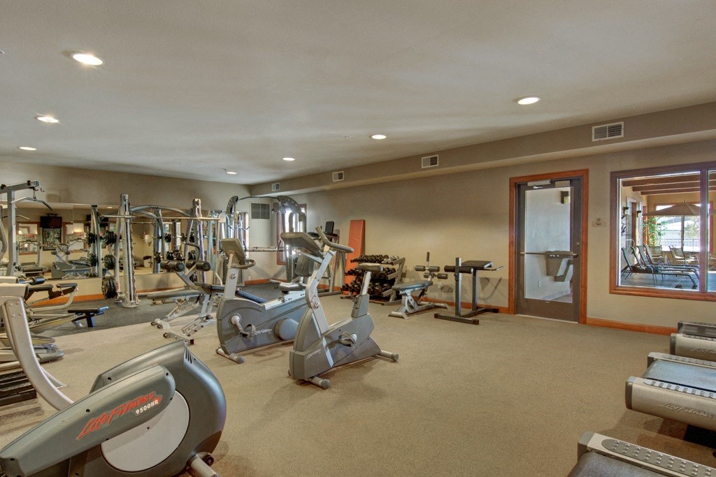 A spacious gym here at Grand River Estates Apartment Homes with various exercise equipment including treadmills, stationary bikes, weights, and mirrors. The room is well-lit and organized, creating an inviting atmosphere.