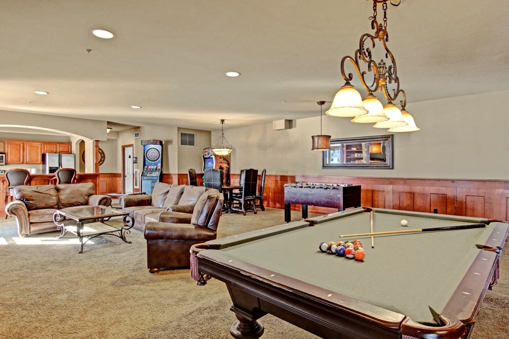 Luxurious game room here at Grand River Estates Apartment Homes featuring a pool table with cues and balls, leather sofas, foosball table, dartboard, arcade game, and warm ambient lighting.
