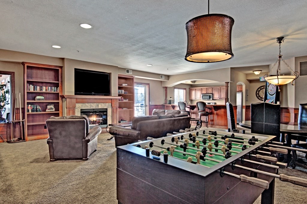 Cozy recreation room here at Grand River Estates Apartment Homes with a foosball table, armchairs by a fireplace, bookshelves, large TV, and bar area. Warm lighting and relaxed atmosphere.