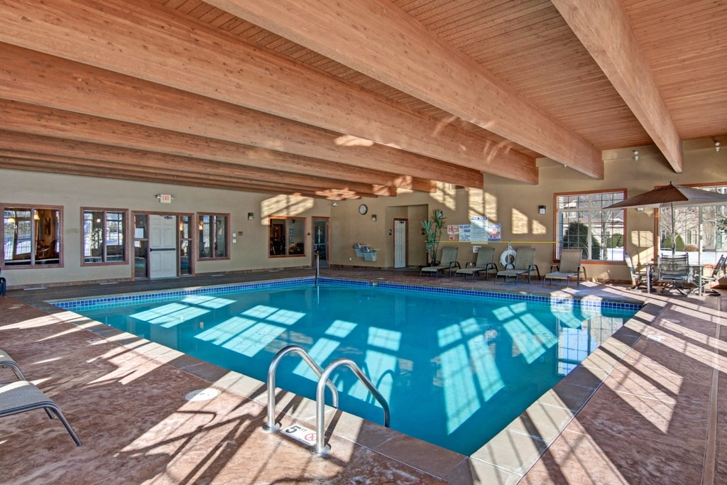 Indoor pool here at Grand River Estates Apartment Homes with clear water and blue tiles, set in a cozy room with wooden beams. Sunlight streams through large windows, casting reflections on the water.