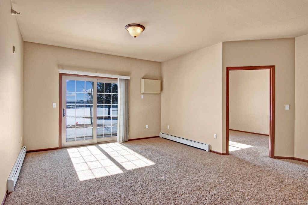 Bright, empty room here at Grand River Estates Apartment Homes with beige walls and carpet, large sliding glass door letting in sunlight, and open doorway connecting to another room. Serene and spacious.