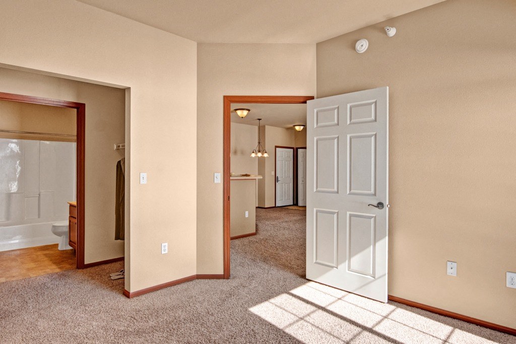 Vacant apartment interior here at Grand River Estates Apartment with beige walls and carpet. Open door reveals dining area with pendant lights. Bathroom entrance on the left, creating a warm, inviting feel.