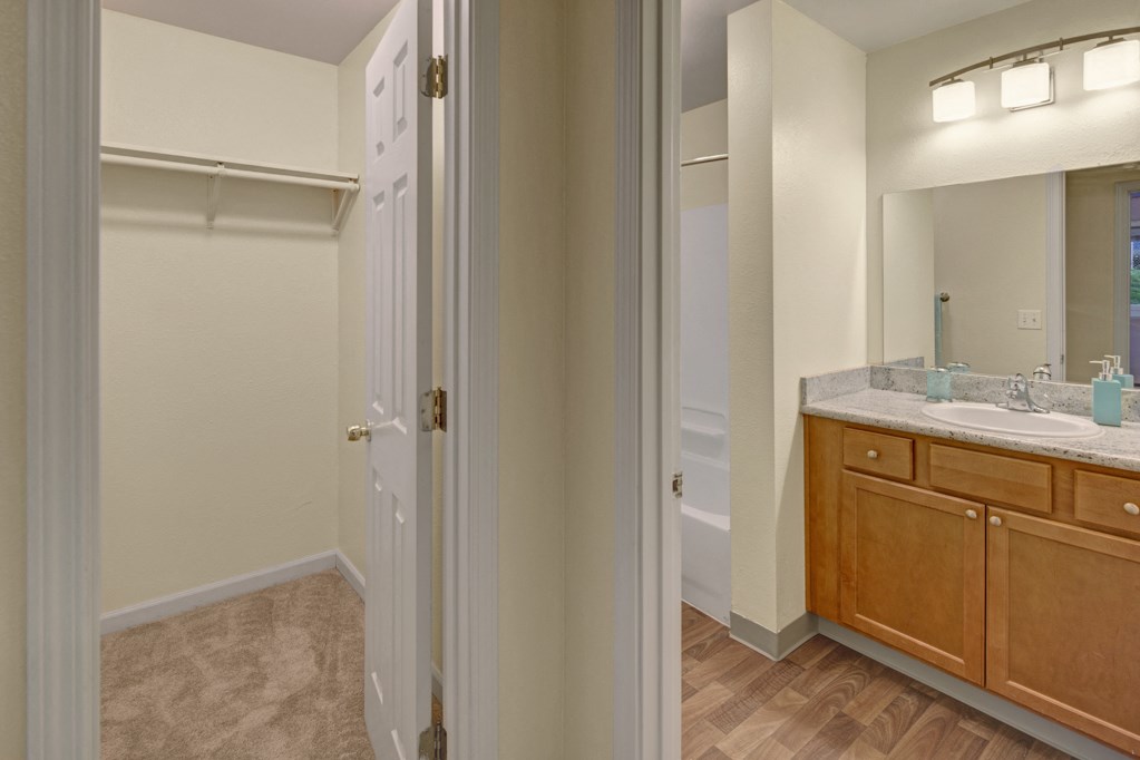 A modern bathroom here at Hillside Chalet Apartments features a spacious vanity with a marble countertop, wooden cabinets, and dual sinks. Adjacent is a carpeted closet space.