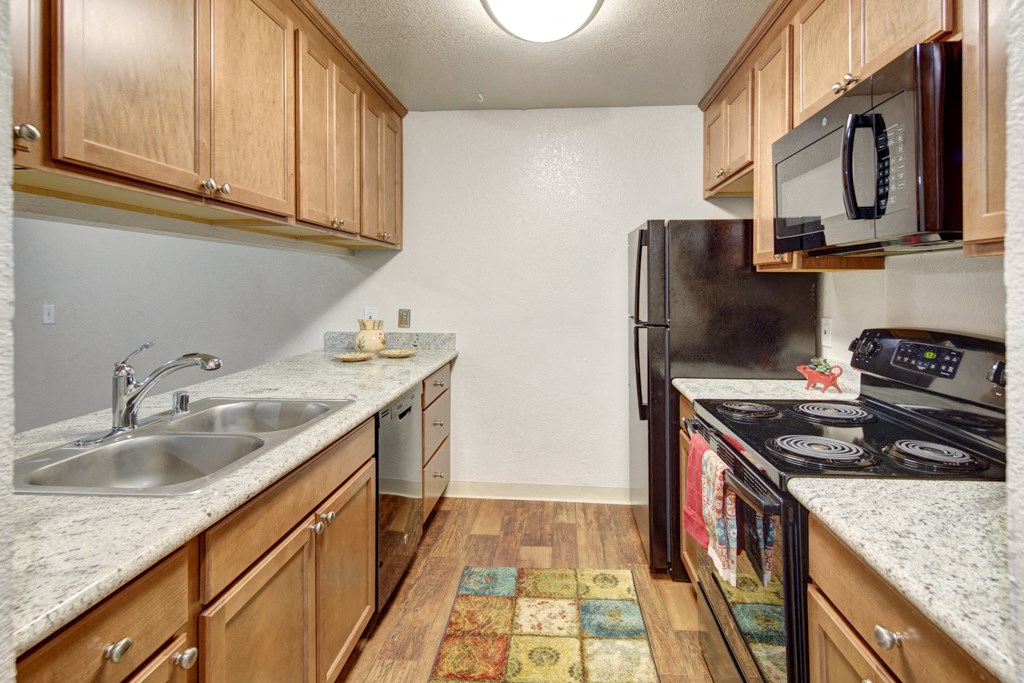 Modern kitchen here at Hillside Chalet Apartments with light wood cabinets, granite countertops, and stainless steel appliances. A colorful rug adds warmth, with an inviting, tidy feel.