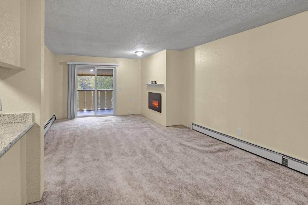 Empty living room here at Hillside Chalet Apartments with beige walls and carpet, featuring a corner electric fireplace. Sliding glass door leads to a balcony with green foliage outside.
