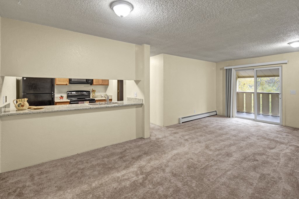 Spacious empty living room here at Hillside Chalet Apartments with beige carpet, open kitchen with light wood cabinets, and large sliding glass doors leading to a balcony with greenery.